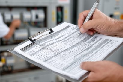 Technician checks electrical system controls and makes notes in a maintenance journal inside a clean, well-lit industrial facility with organized panels.